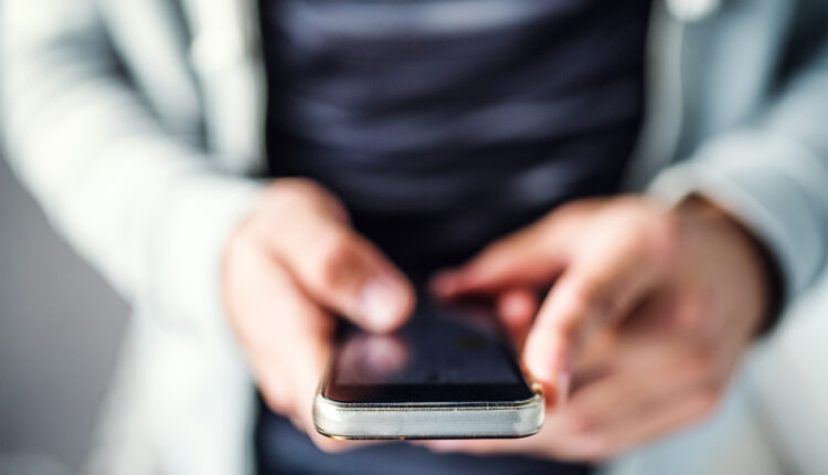 A closeup photo shows a man holding a smartphone in his hands. The shot is tightly focused on his phone, the rest of the photo is blurred.