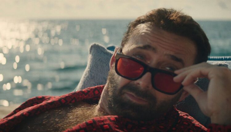 Nic Cage lowers his red sunglasses by the water in The Unbearable Weight of Massive Talent